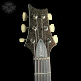 Close-up of an electric guitar headstock with a dark glitter finish, cream tuning pegs, and a silver 'Custom' script on the neck plate, shown against a black background with a Tone Shop watermark.