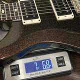 A close-up of a black glitter electric guitar body resting on a digital kitchen scale displaying 7.68 lbs (3.