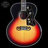 A close-up studio shot of an acoustic guitar body against a black background, showing a sunburst finish that transitions from bright yellow-orange in the center to deep red and black around the edges. The guitar features an ornate black pickguard with gold and white floral details, a white binding, and a dark fretboard.