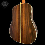 Close-up of the back of an acoustic guitar showing a rich dark brown wood grain finish with a light cream binding, set against a solid black background. A Tone Shop watermark is visible in the top left corner.