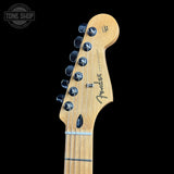 Close-up of the headstock of a Fender Stratocaster electric guitar, showing the natural maple wood neck and six black tuning pegs against a solid black background.