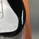 Close-up detail of a glossy black electric guitar body corner with a white pickguard and part of a wood neck or stand visible.