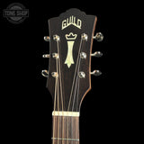 Close-up of a Guild acoustic guitar headstock and upper neck, showing a dark wood finish, gold-toned logo and tuning pegs, and six strings against a black background.