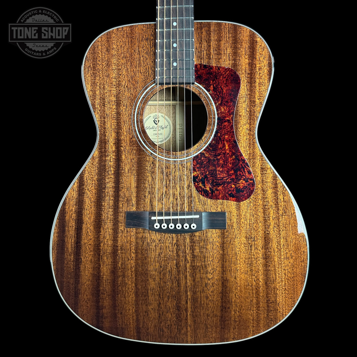 A close-up studio shot of an acoustic guitar body against a black background, showing a natural wood grain finish, a red tortoiseshell-style pickguard, and part of the neck. A Tone Shop watermark is visible in the top left corner.
