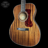 A close-up studio shot of an acoustic guitar body showing a rich wood grain finish, a dark red flame-pattern pickguard, and a black bridge, set against a solid black background with a Tone Shop watermark in the top left corner.