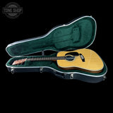 An acoustic guitar with a natural wood finish resting inside an open black hard case with a green velvet interior, shown against a solid black background. A Tone Shop watermark is visible in the top-left corner.