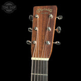 Close-up of the headstock of a Martin & Co acoustic guitar against a solid black background, showing the brand name in gold script on a dark wood headstock with six silver tuning pegs.
