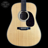 A close-up studio shot of an acoustic guitar body against a solid black background, featuring a natural wood finish, a dark tortoiseshell pickguard, and a black bridge. A circular 'TONE SHOP' watermark is visible in the top left corner.