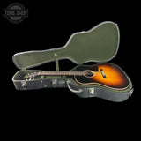 An acoustic guitar with a sunburst finish resting inside an open hard guitar case against a solid black background. A Tone Shop watermark is visible in the top-left corner.