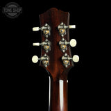 Close-up of the headstock of a dark brown wooden acoustic guitar against a solid black background, showing six tuning pegs and a Tone Shop watermark in the top left corner.