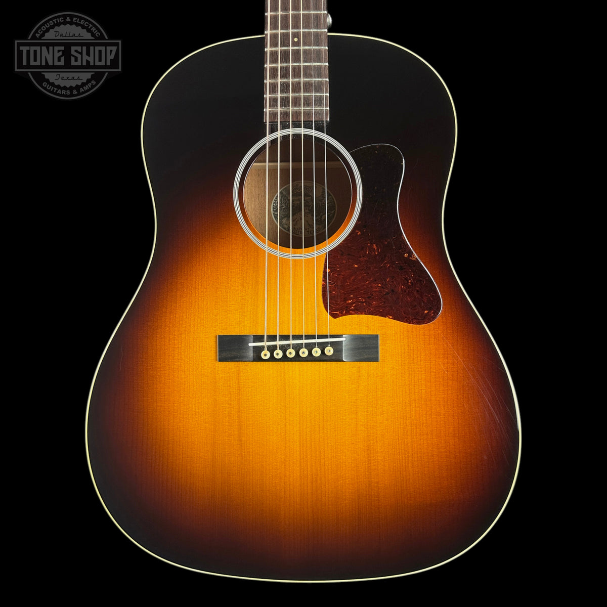 A classic acoustic guitar with a sunburst finish, featuring a bright orange center fading to a dark black edge. The guitar is shown from a front-facing angle against a solid black background, with a Tone Shop watermark in the upper left corner.