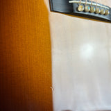 Close-up detail of a guitar body showing the orange wood fingerboard, white fabric cover, and part of the black bridge with metal strings.