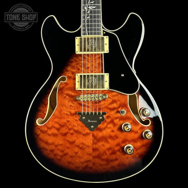 An electric guitar with a vibrant orange-to-brown flamed maple top, black pickguard, and gold hardware, shown against a solid black background.