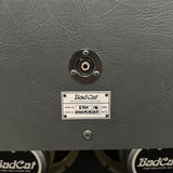 Close-up of the rear panel of a Bad Cat tube amplifier, showing a textured dark gray metal surface, a circular connector, and a metal model plate with handwritten ownership information.