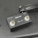 A BadCat Era 30 guitar effects pedal with a black metal casing, two silver footswitches, and white printed text, resting on a black textured leather surface.