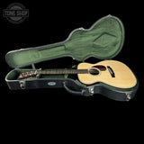An acoustic guitar with a natural wood finish resting inside an open black hard case with green padding, shown against a solid black background. A 'TONE SHOP' watermark is visible in the top-left corner.