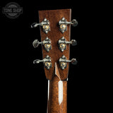 Close-up of the headstock of an acoustic guitar, showing six tuning pegs with decorative gold-toned centers and a glossy brown wood finish, set against a solid black background.