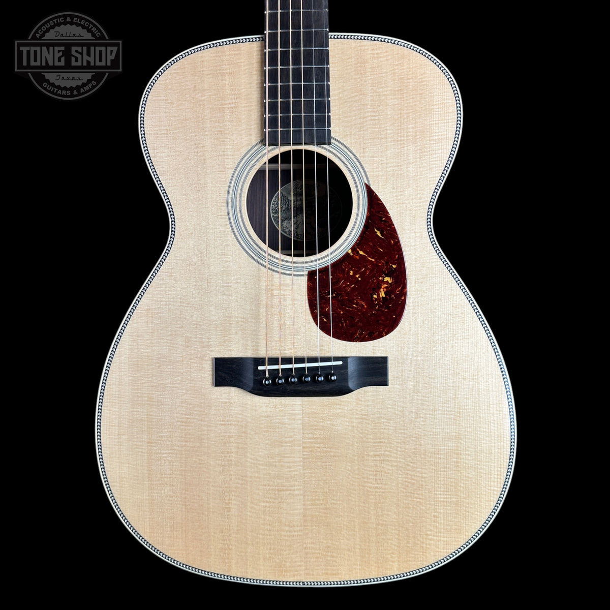 A close-up studio shot of an acoustic guitar body against a solid black background, featuring a natural flamed wood top, a dark reddish-brown pickguard, and a black bridge. A Tone Shop watermark is visible in the top left corner.