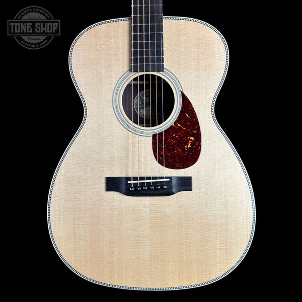 A close-up studio shot of an acoustic guitar body against a solid black background, featuring a natural flamed wood top, a dark reddish-brown pickguard, and a black bridge. A Tone Shop watermark is visible in the top left corner.
