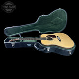 An acoustic guitar with a natural wood top and dark neck, resting inside an open black hard case with a green padded interior, shown against a solid black background. A Tone Shop watermark is visible in the top-left corner.