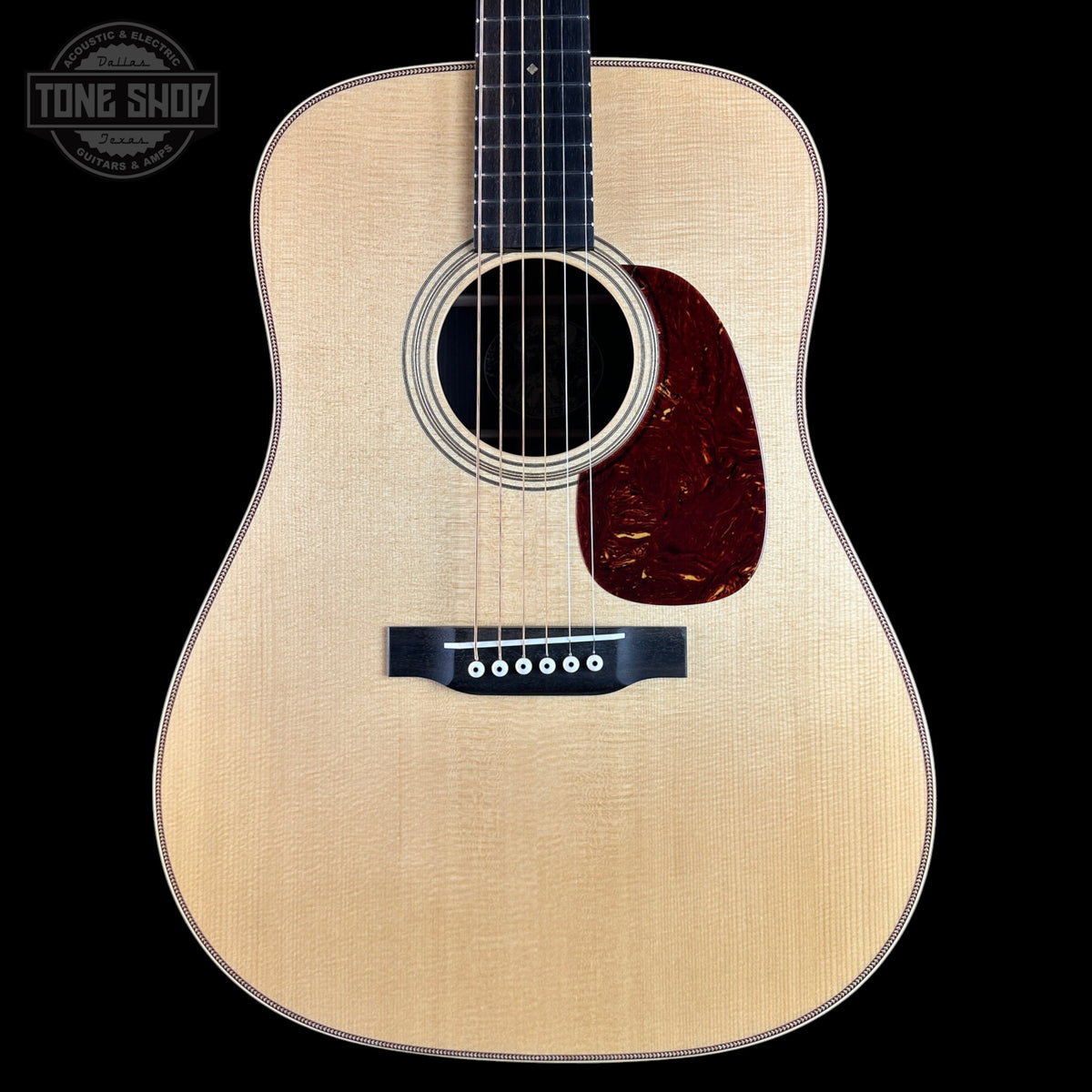 A front-facing acoustic guitar with a natural wood finish and a dark reddish-brown pickguard, shown against a solid black background with a Tone Shop watermark in the top-left corner.