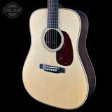 A close-up of an acoustic guitar body with a natural wood top, dark brown sides, and a dark red pickguard, shown against a black background with a Tone Shop watermark in the top left.