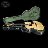 A natural wood acoustic guitar resting inside an open black hard case with a green velvet-lined interior, photographed on a solid black background. A Tone Shop watermark is visible in the top-left corner.