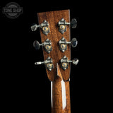 Close-up of the headstock of a six-string acoustic guitar, showing six tuning pegs with gold and silver accents on a dark brown wood finish, set against a solid black background.
