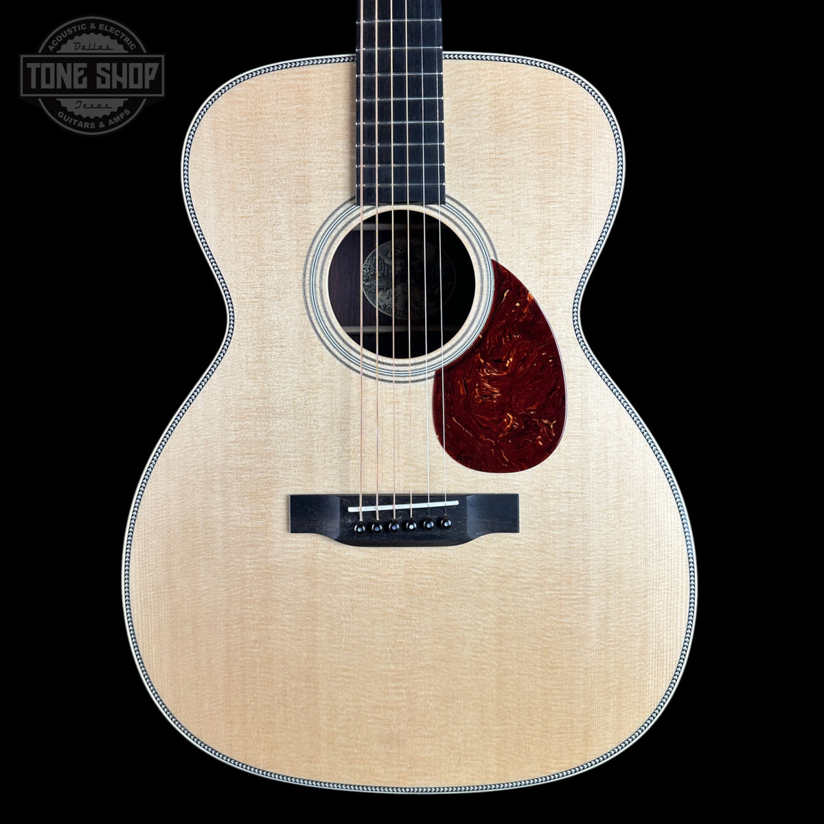 An acoustic guitar with a natural wood top, dark fretboard, and a dark red carved pickguard, shown against a solid black background with a Tone Shop watermark in the top-left corner.