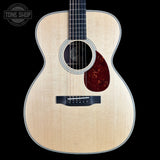 An acoustic guitar with a natural wood top, dark fretboard, and a dark red carved pickguard, shown against a solid black background with a Tone Shop watermark in the top-left corner.