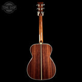 The back view of an acoustic guitar with a rich dark brown wood grain finish, shown against a solid black background with a Tone Shop watermark in the top left corner.