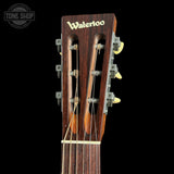 Close-up of the headstock of a wooden acoustic guitar on a black background, featuring the 'Waterloo' brand name in gold lettering and six tuning pegs.