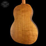 The back of a wooden acoustic guitar, showcasing a rich warm brown wood grain finish, set against a solid black background with a Tone Shop watermark in the top left corner.