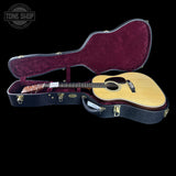 An acoustic guitar with a natural wood finish sits inside an open black hard case with burgundy velvet lining, photographed against a solid black background. A Tone Shop watermark is visible in the top-left corner.