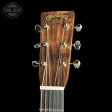 Close-up of a Martin & Co acoustic guitar headstock, showing the brand name in gold script on a dark brown wood background, with six silver tuning pegs and strings against a black background.