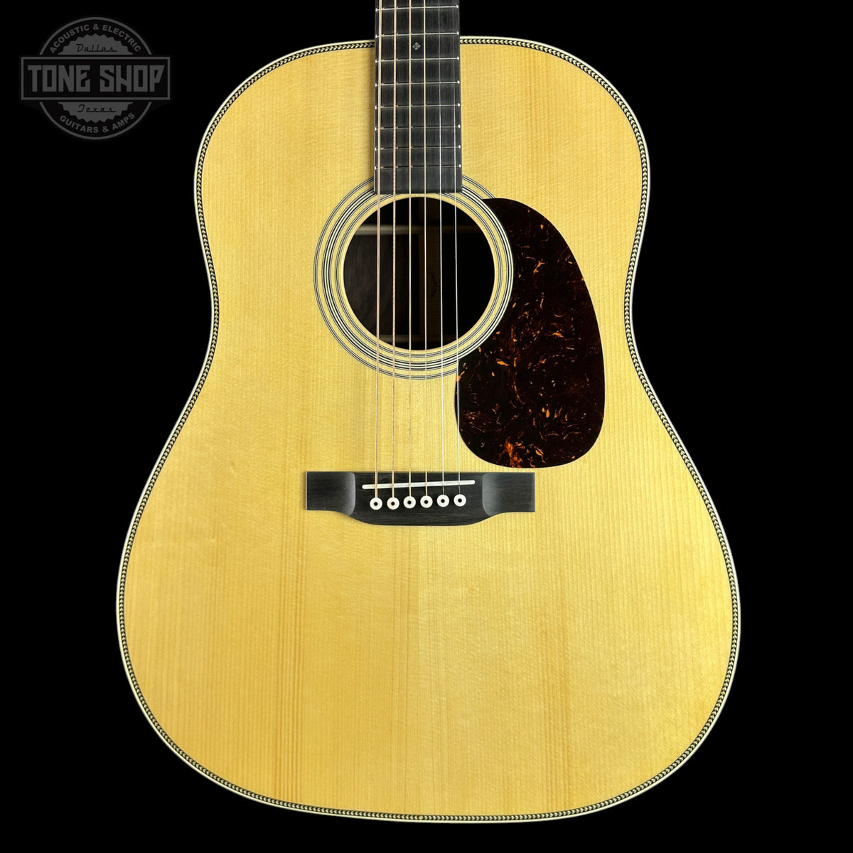 A close-up studio shot of an acoustic guitar body against a solid black background, featuring a natural wood finish, a dark tortoiseshell pickguard, and a white beaded edge.