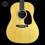 A close-up studio shot of an acoustic guitar body against a solid black background, featuring a natural wood finish, a dark tortoiseshell pickguard, and a white beaded edge.