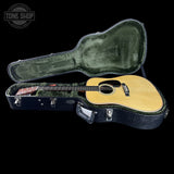 An acoustic guitar with a natural wood body and dark neck, resting inside an open black hard case with green plush lining, shown against a solid black background. A Tone Shop watermark is visible in the top-left corner.