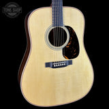 A close-up studio shot of an acoustic guitar body against a solid black background, featuring a natural wood top, dark sides, and a tortoiseshell-style pickguard.