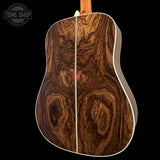 The back of an acoustic guitar featuring a rich, dark brown wood grain pattern with swirling natural patterns, a light-colored center seam, and white binding, shown against a solid black background with a Tone Shop watermark in the top left.