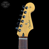 Close-up of a Fender Stratocaster electric guitar headstock in a natural maple finish, showing six black tuning pegs and the Fender logo, set against a solid black background.