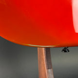 Close-up detail of a glossy bright red chair seat or backrest resting on a wooden stand, with a small black metal support visible against a dark gray background.