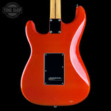Close-up of the back of a glossy orange electric guitar body with a black pickguard, black hardware, and part of a light wood neck visible against a black background. A Tone Shop watermark appears in the upper left corner.