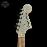 Close-up of the headstock of a Squier Jaguar electric guitar against a solid black background, showing the silver glitter finish, six tuning pegs, and the Squier Jaguar branding.