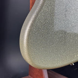 Close-up of a guitar body edge with a metallic glitter finish in a muted olive-silver tone, mounted on a wooden stand against a dark background.