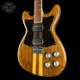 A classic electric guitar with a natural wood finish, featuring a dark brown pickguard and a vertical light wood stripe down the center. The guitar is photographed against a solid black background with a Tone Shop logo in the top-left corner.
