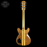 The back view of an electric guitar with a natural wood body and maple neck, shown against a solid black background with a Tone Shop watermark in the top left corner.