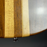 Close-up of a curved wooden surface with vertical striped wood grain in light tan, medium brown, and dark brown tones, with a small metallic screw visible near the bottom against a dark background.