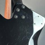 Close-up of the back of a black electric guitar body with visible scratches, silver screws, a chrome hardware plate, and part of the wooden neck at the top left, photographed against a neutral gray background.