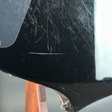 Close-up of a glossy black guitar body showing multiple scratches and scuffs on the finish, with part of a wooden stand visible below.